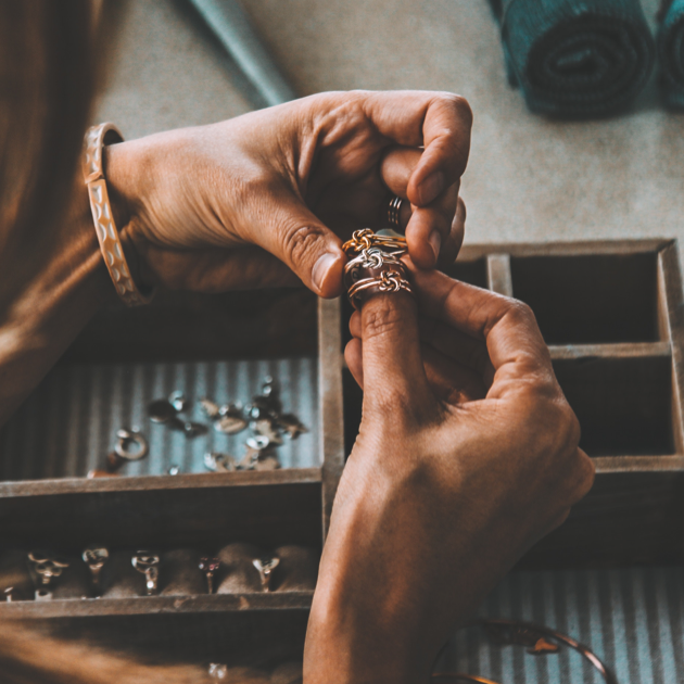 A jeweler at their worktable with gold, silver and rose gold rings stacked on their right thumb.