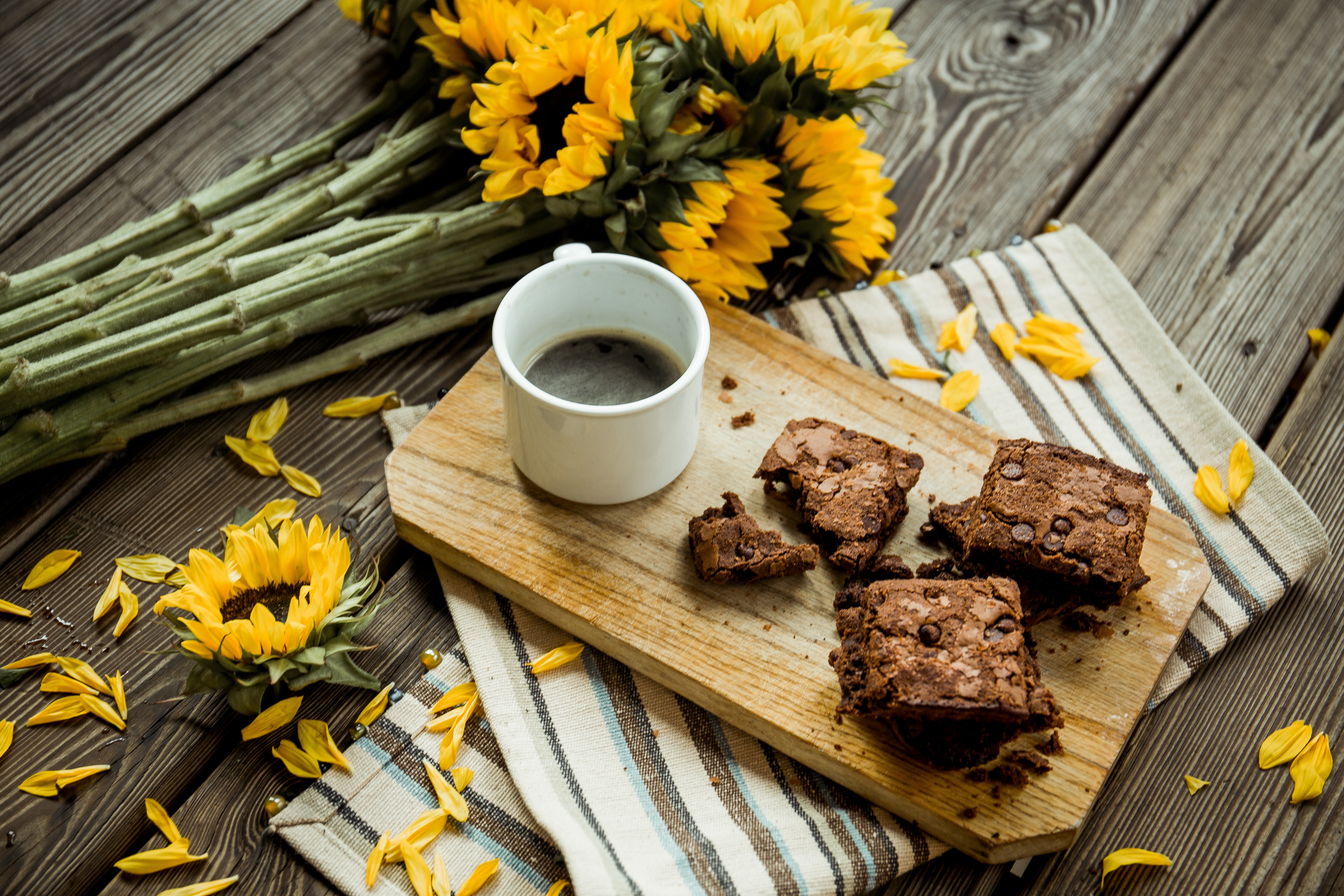 A few brownie squares on a cutting board next to a cup of coffee and a bouquet of fresh sunflowers.